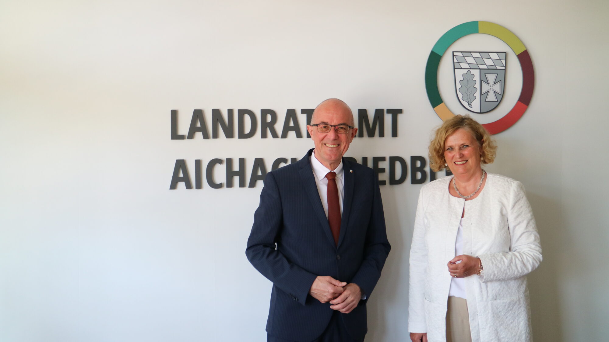 Landrat Dr. Klaus Metzger (links) und Regierungspräsidentin Barbara Schrettler (rechts) freuten sich auf ihr Kennenlernen und stehen hier auf dem Bild vor dem Landratsamtslogo.