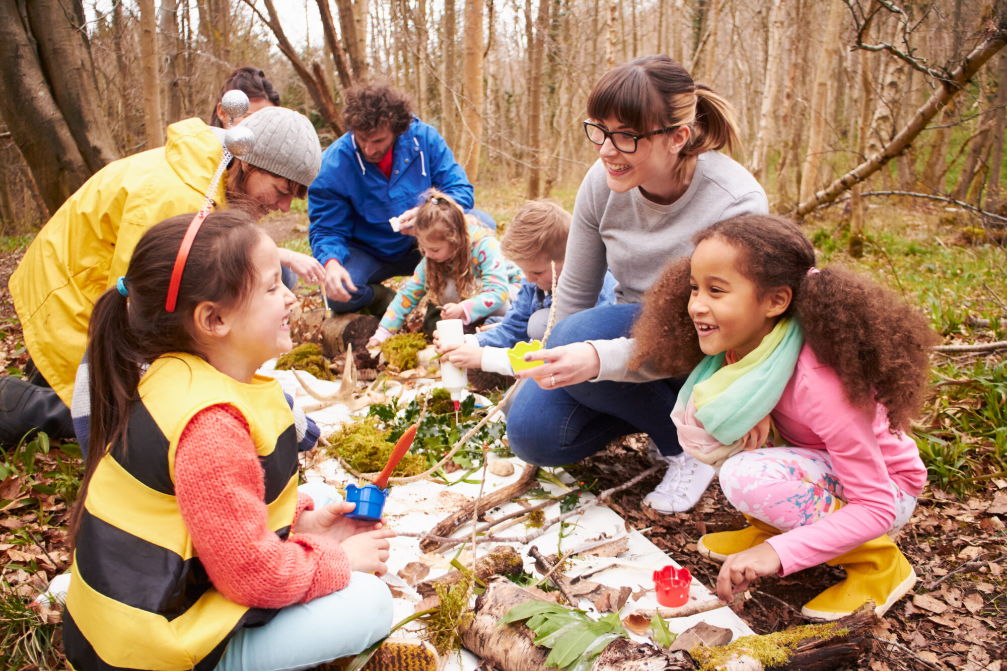Hier sehen Sie Kinder mit Erwachsenen im Wald. Vor ihnen auf dem Boden verschiedene Naturmaterialien