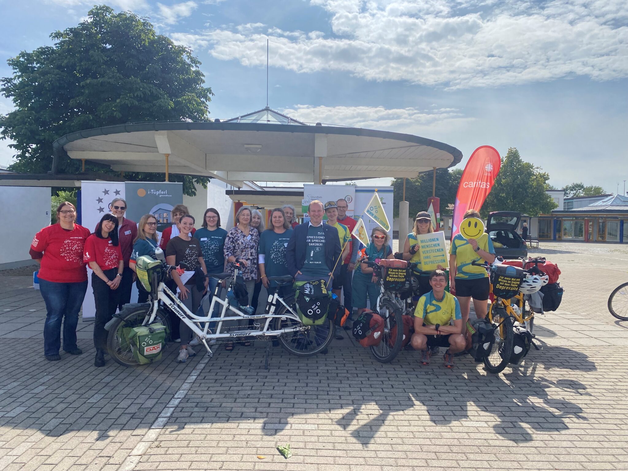 Hier sieht man eine Gruppe von ca. 20 Personen auf einem Pausenhof zusammen stehen und in die Kamera lachen. Im Hintergrund sieht man noch einige Roll-ups der Organisationen. Außerdem noch Tandemfahrräder. Das Wetter ist schön. Die Personen haben bunte Tshirts an auf denen steht MUT-Tour 2025: Depressionen, wir sprechen darüber